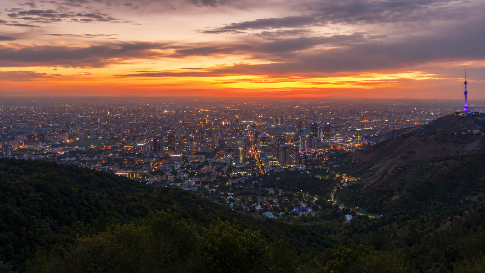 Sunset view from Kok-Tobe over Almaty with city grid, mountains, and wide horizon.