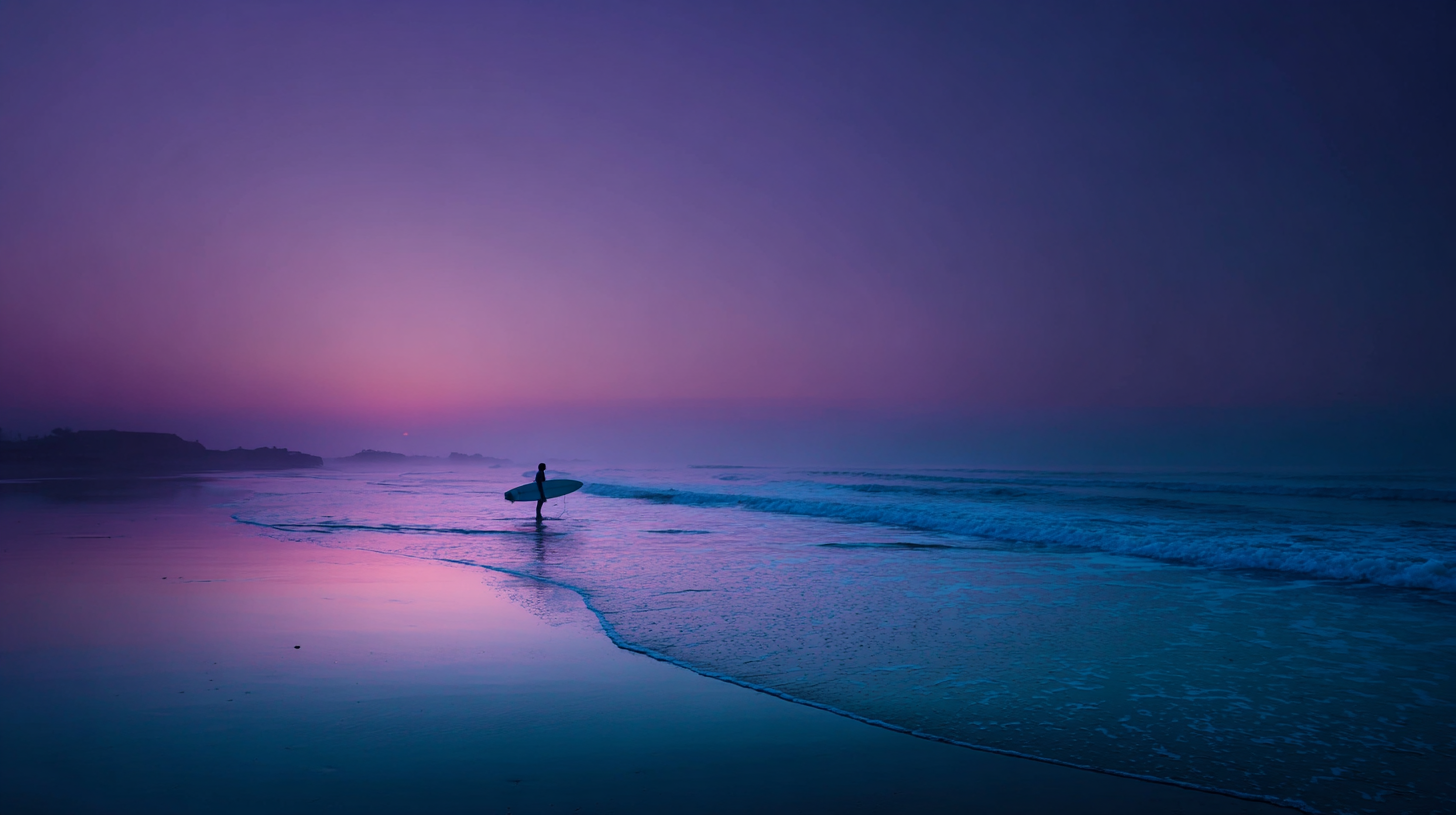 Surfer standing at water’s edge at twilight, reflecting.